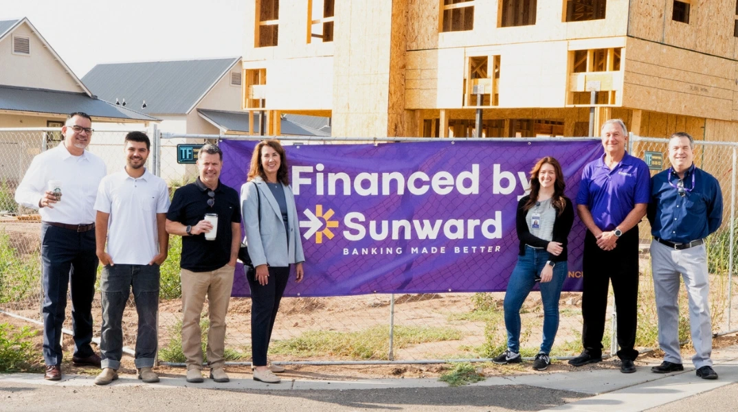 Sunward leadership and employees standing in front of a fence that has a "Financed by Sunward" banner on it. Behind the fence is one house that is being constructed and several others that are complete.