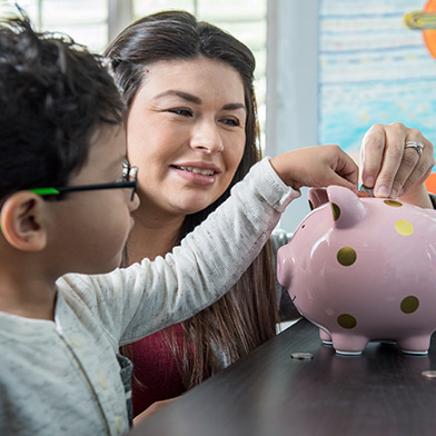 Mom helping young boy insert coins into a piggy bank.