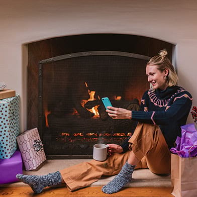 Woman using cell phone by fireplace with wrapped gifts