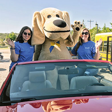 Sandy the Lab sitting in a convertible auto posing with two credit union employees.