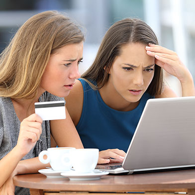 Two young women frowning as they look at a computer screen; one is holding a credit or debit card in her hand.